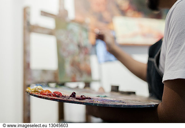 Midsection of painter holding palette while painting in studio