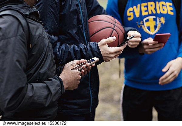 Midsection of phone addicted friends wearing warm clothing while standing on basketball court