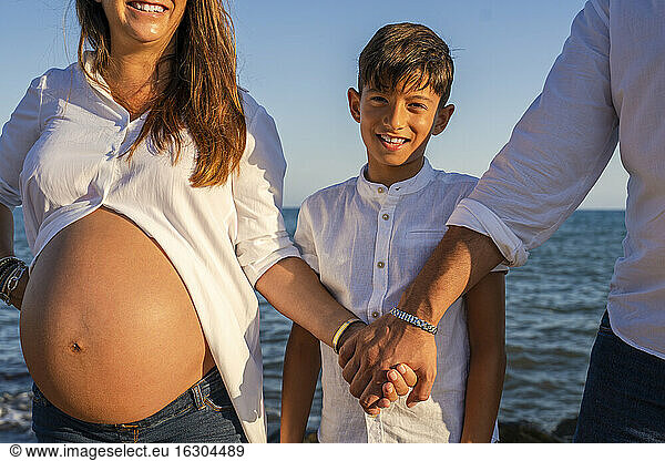 Midsection of pregnant woman holding hand of man while son standing in between against beach