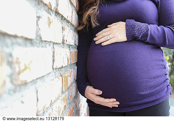 Midsection of pregnant woman touching belly while standing by brick wall