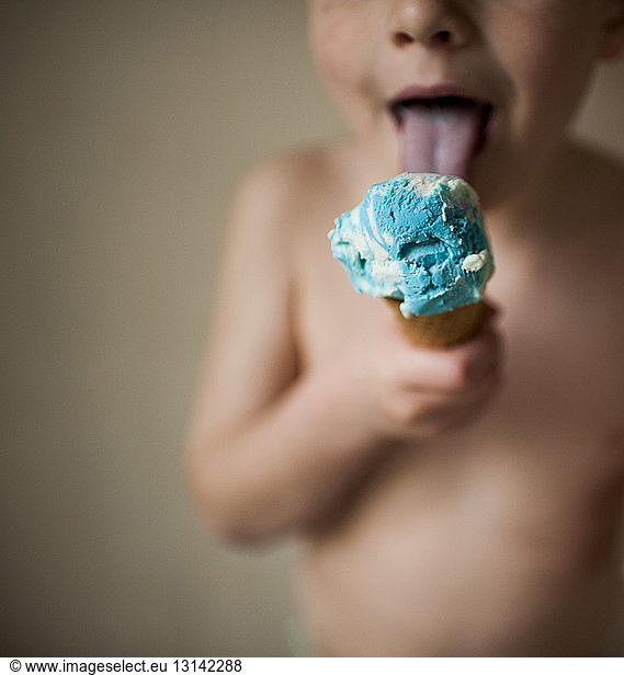 Midsection of shirtless boy licking ice cream while standing against wall at home