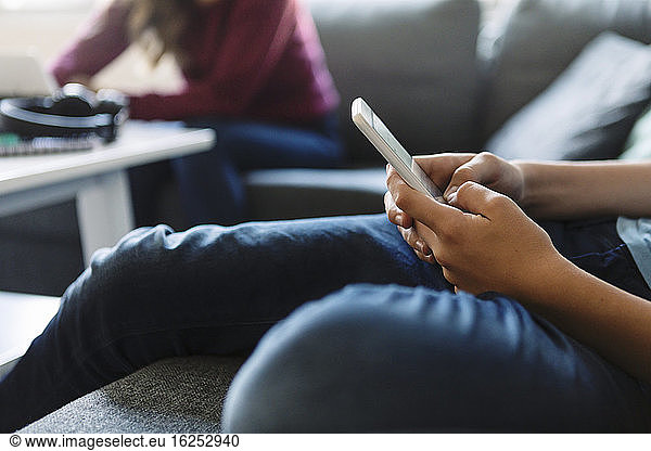 Midsection of teenager using phone while sister sitting on sofa at home