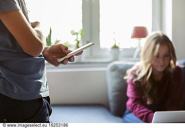 Midsection of teenager using technology while sister sitting on sofa at home