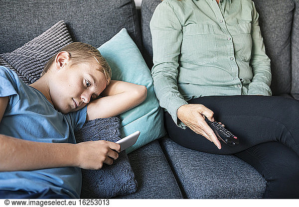 Midsection of woman holding remote control while son using phone on sofa