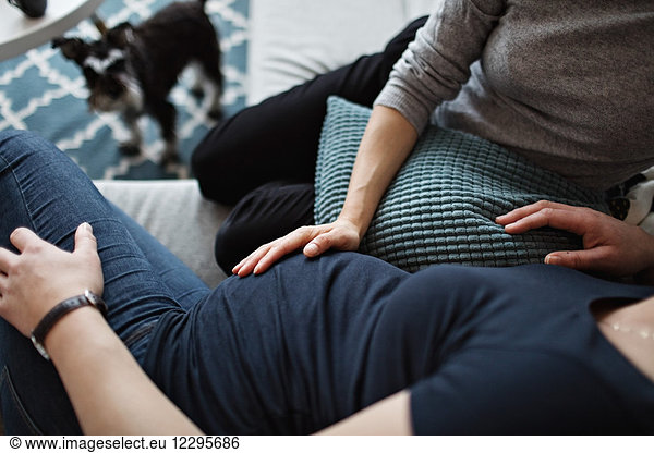 Midsection of woman touching girlfriend's abdomen while sitting on sofa at home