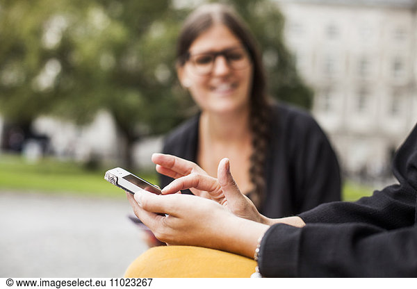 Midsection of woman using smart phone sitting with friend outdoors
