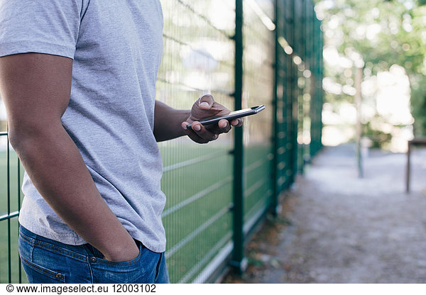 Midsection of young man standing against fence while using smart phone