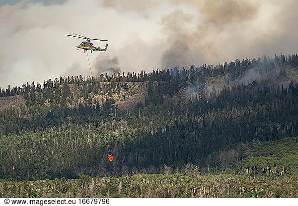 Military helicopter flying with fire retardant while smoke emitting from forest