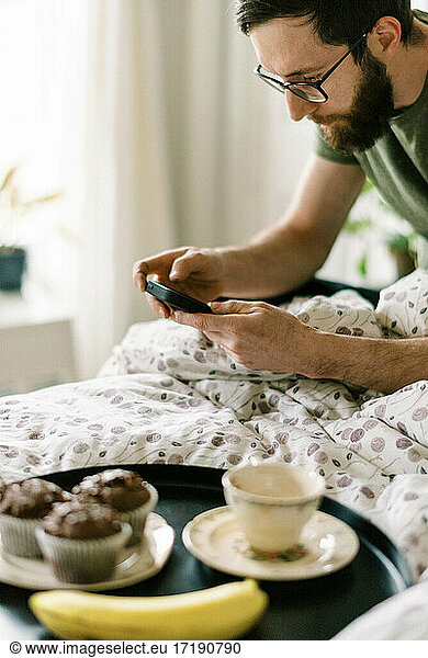 Millennial man having breakfast and tea in bed while scrolling phone