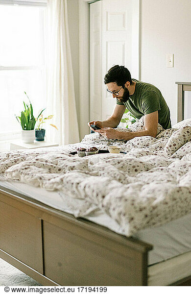 Millennial man having breakfast and tea in bed while scrolling phone