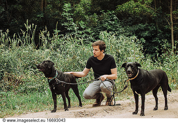 Millennial-Mann sitzt mit seinen zwei schwarzen Hunden auf der Hut im Wald