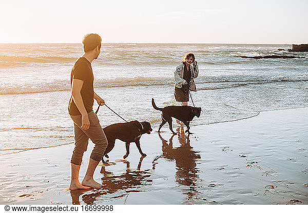 Millennial Mann und Frau mit zwei Hunden zu Fuß am Strand bei Sonnenuntergang