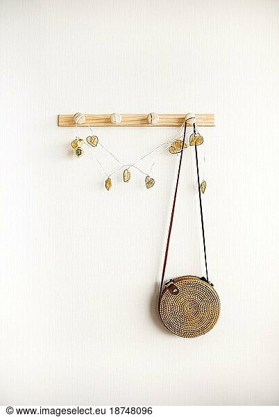 Minimalistic image of round rattan boho crossbody bag hanging on wooden hanger against white wall. Fashion and bohemian style accessories concept. Minimal home interior design