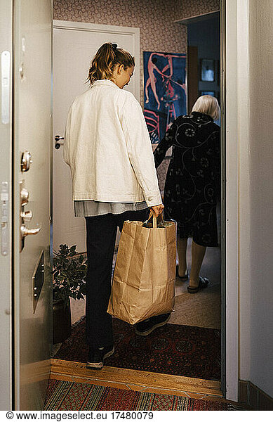 Mitarbeiterin im Gesundheitswesen mit Einkaufstasche betritt Haus hinter älterer Frau