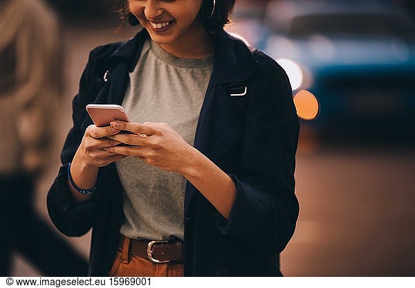 Mitschnitt einer lächelnden jungen Frau  die soziale Medien am Telefon nutzt  während sie in der Stadt steht