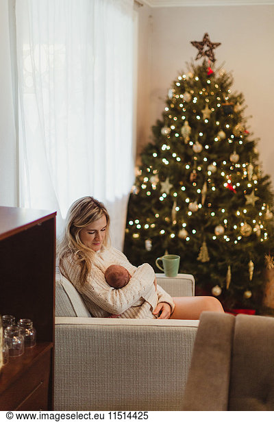 Mittelgroße erwachsene Frau sitzt auf dem Sofa und wiegt ein neugeborenes Baby  das zu Weihnachten in eine Strickjacke gehüllt ist