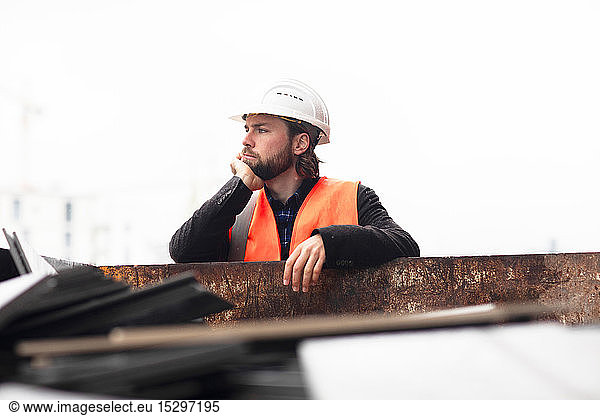 Mittelgroßer erwachsener männlicher Bauarbeiter lehnt an Abfallcontainer lehnend nachdenkend