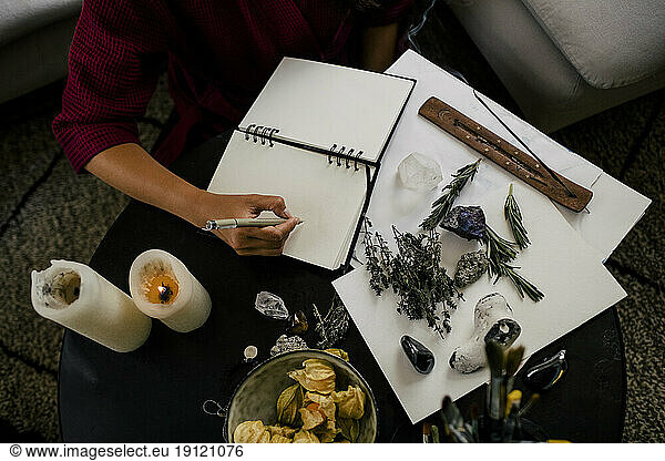 Mittelteil einer Frau  die neben Kräutern und Kristallen auf einem Tisch in ein Buch schreibt