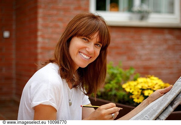 Mittlere erwachsene Frau  die im Freien sitzt und Kreuzworträtsel macht  in die Kamera schaut und lächelt.