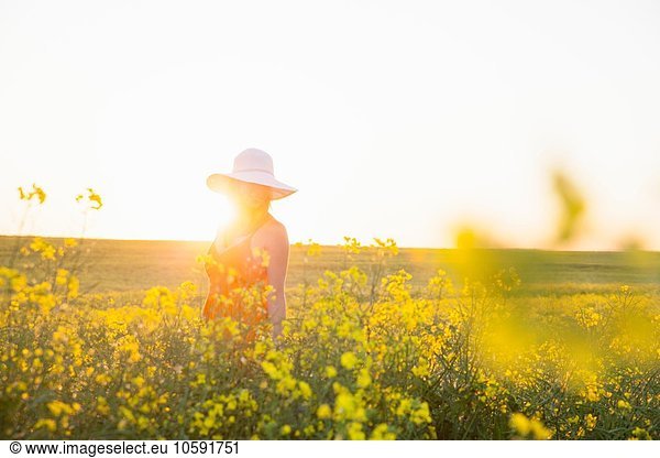 Mittlere erwachsene Frau im Rapsfeld mit Sonnenhut lächelnd  Fokus auf Hintergrund  Linsenflackern