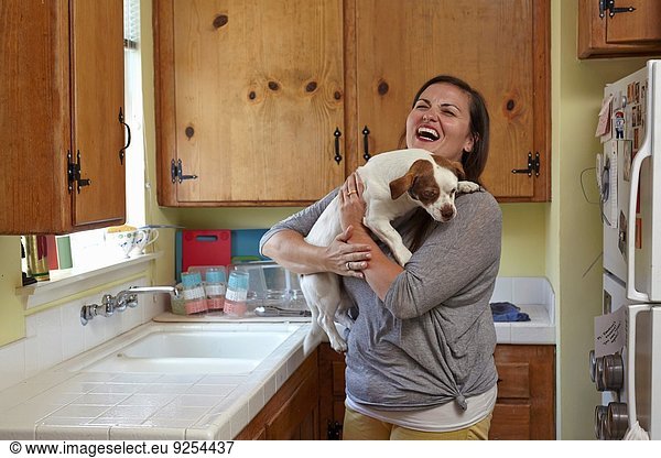 Mittlere erwachsene Frau trägt und streichelt Hund in der Küche