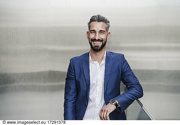 Mittlerer erwachsener Geschäftsmann  der sich lächelnd auf ein Geländer vor einer silberfarbenen Wand stützt
