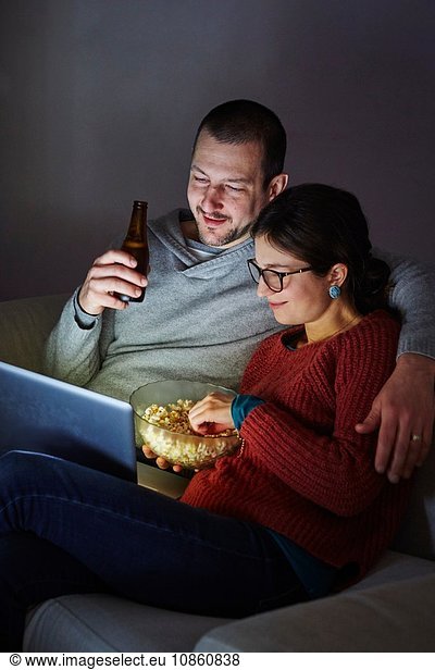 Mittleres erwachsenes Paar sitzt abends auf dem Sofa  isst Popcorn und schaut auf den Laptop