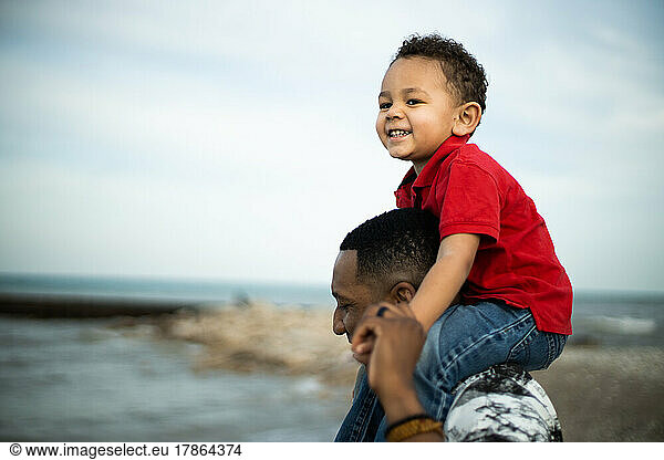 Mixed race son smiles happily on black fathers shoulders on beach