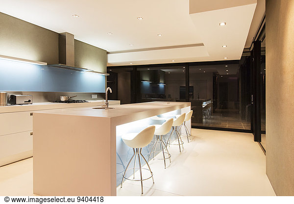 Modern white kitchen with kitchen island and stools illuminated at night