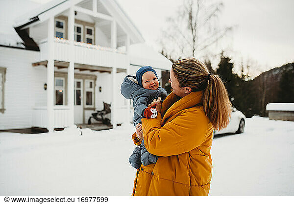 Mom and adorable baby smiling on cold snowy day outside white house