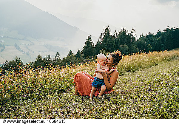 Mom kissing her baby sitting on the grass in the Alps
