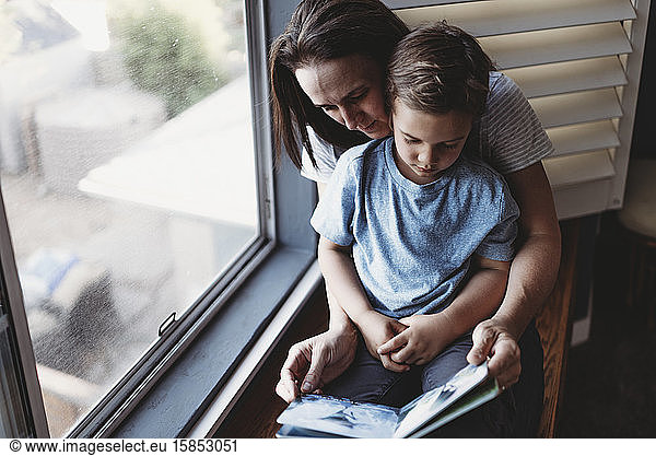 Mom reading book with son on her lap next to window