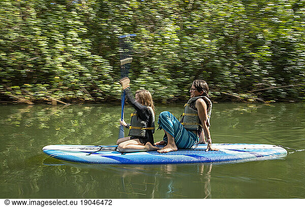 Mom sits behind daughter on paddle board on tropical river