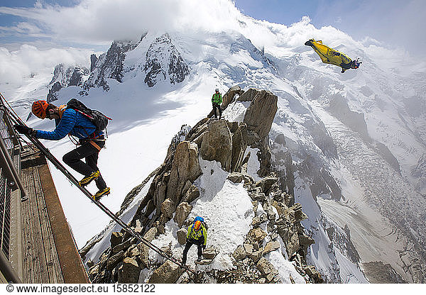 Mont-Blanc von der Aiguille Du Midi oberhalb von Chamonix  Frankreich  mit Bergsteigern auf der Cosmiques Arete  die die Leiter zur Seilbahnstation erklimmen  und einem Base Jumper in einer Wing-Suite beim Fliegen