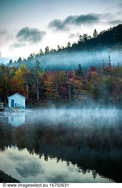 Moody autumn morning with fog rising above calm lake in the mountains.