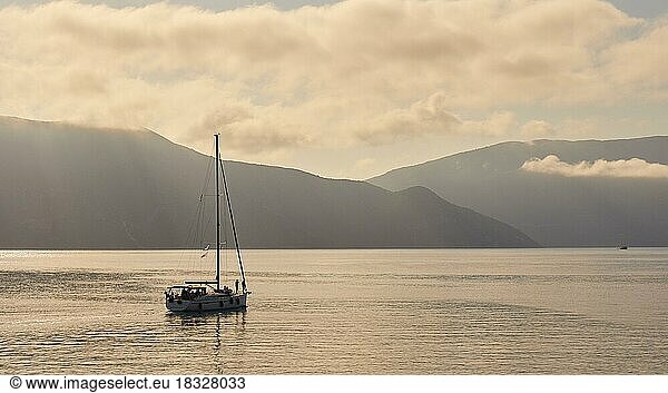 Morgenlicht  Hafeneinfahrt  fahrendes Segelboot ohne Segel  Sonnenstrahlen  Wolken  glattes Meer  Ithaka  Hafenort  Fiskardo  Insel Kefalonia  Ionische Inseln  Griechenland  Europa