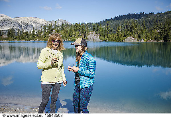 Mother and daughter enjoy a glass of wine by a mountain lake.