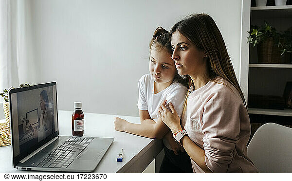 Mother and daughter listening to pediatrician over video call on laptop at home