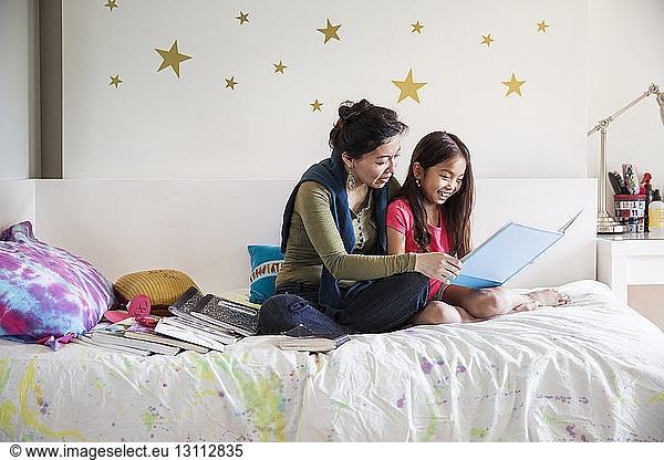 Mother and daughter reading book while sitting on bed at home