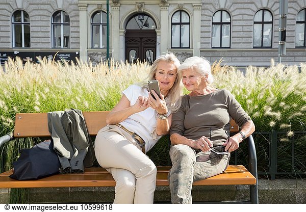 Mother and daughter sitting on bench together  looking at smartphone  outdoors