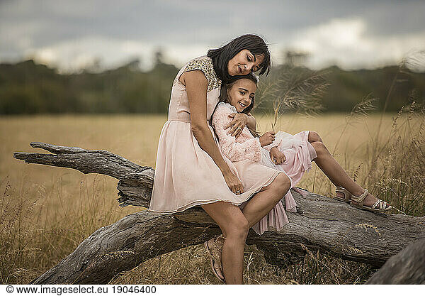 mother and daughter sitting on tree branch in open field hugging