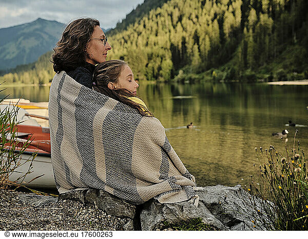 Mother and daughter wrapped in blanket sitting at lakeshore
