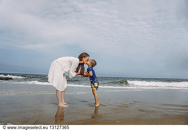 Mother and son kissing on mouth while standing on shore at beach against sky