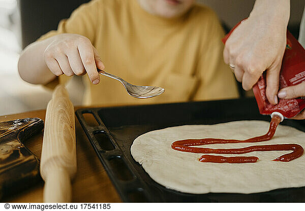 Mother and son spreading tomato sauce on dough in tray at home