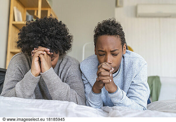 Mother and son with eyes closed praying by bed at home