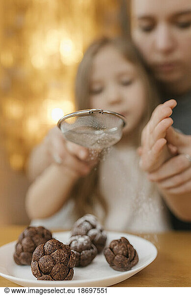 Mother assisting daughter sifting powdered sugar on chocolate pine cones