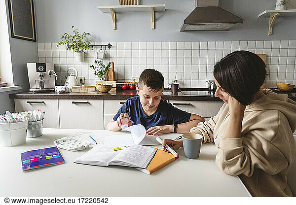 Mother assisting son in homework while sitting at dining table in kitchen at home