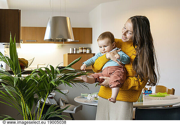 Mother carrying baby boy playing with houseplant at home