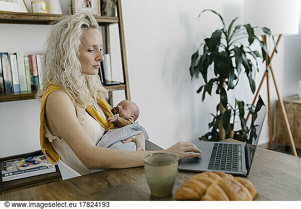 Mother carrying baby in arm using laptop at home