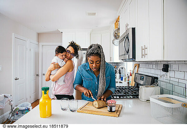 Mother carrying cute son while girlfriend preparing breakfast in kitchen at home